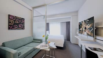 Habitación de hotel con cama, sofá, mesita con revista y flores, televisor y obras de arte en las paredes.