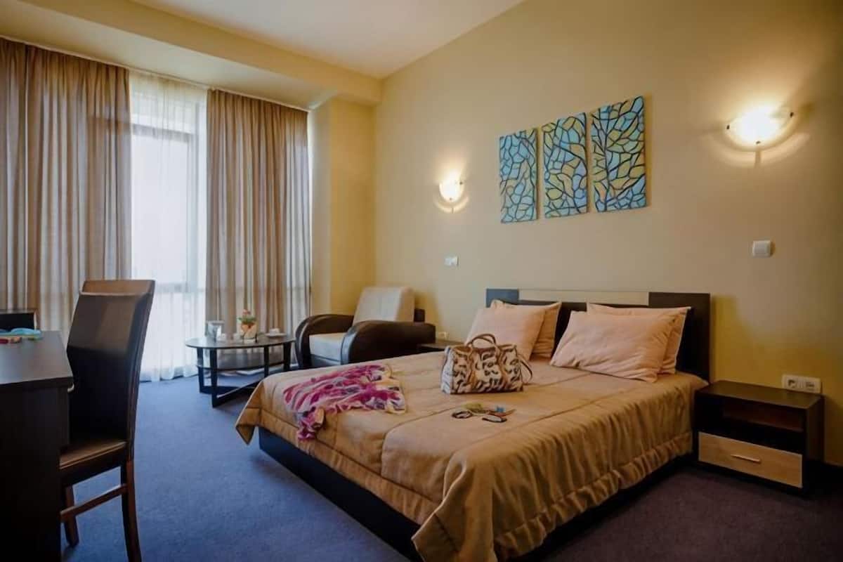 A hotel room with a bed, a desk, a chair, a nightstand, and a wall-mounted lamp.