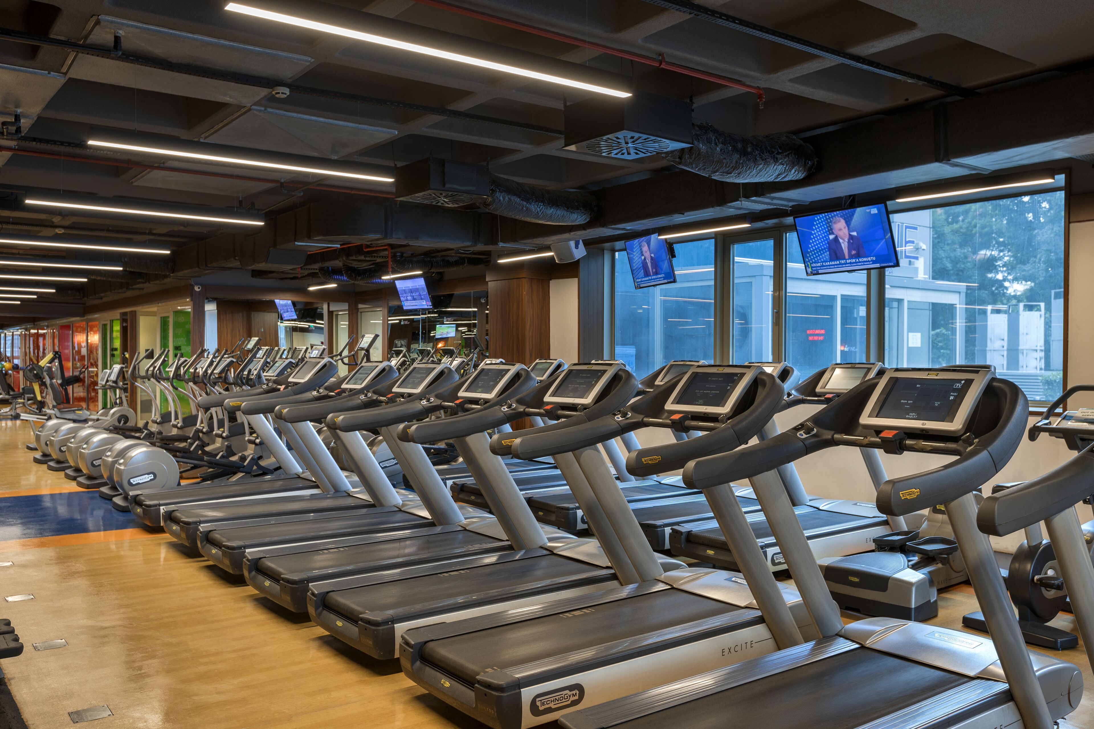 A row of treadmills in a gym with large windows and multiple screens displaying news.