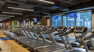 A row of treadmills in a gym with large windows and multiple screens displaying news.