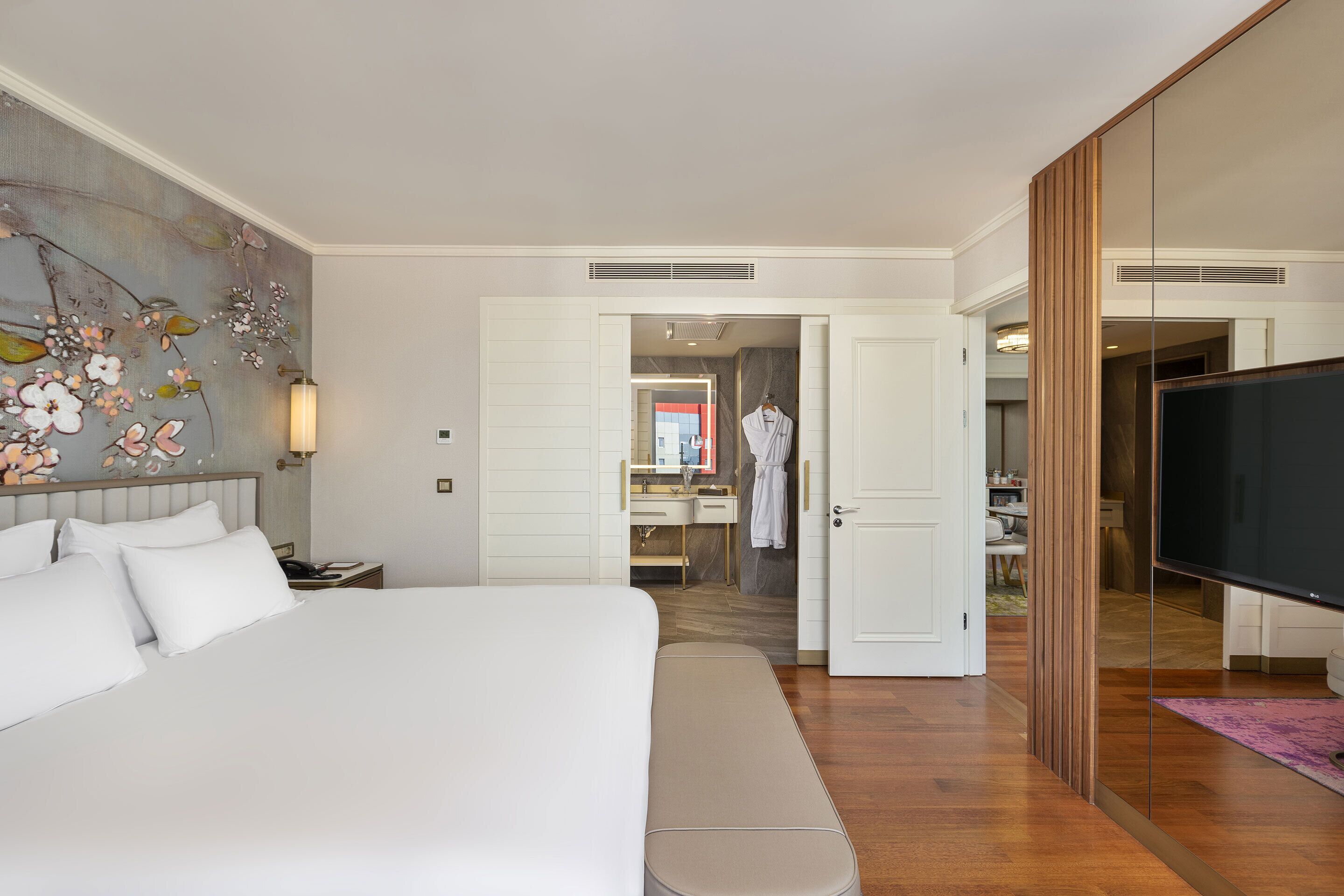 A hotel room with a large bed, a bedside table, a mirror, a wardrobe, a television, and a bathroom visible through an open door.