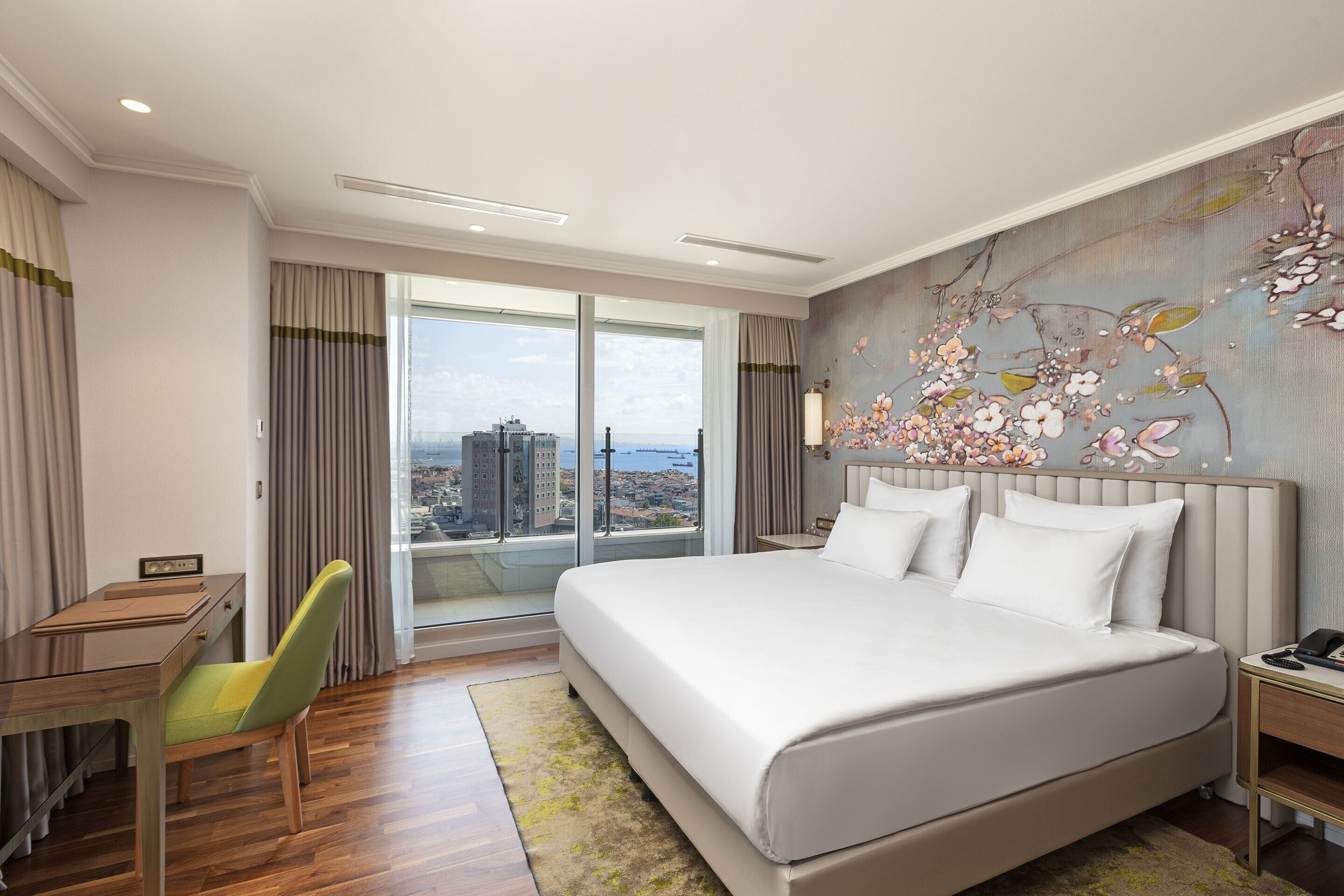 A modern hotel room with a large bed, a desk, and a chair. There is a view of the city through the windows.
