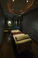 A spa room with two beds, a wooden floor, a glass wall with plants, and a ceiling with recessed lighting.