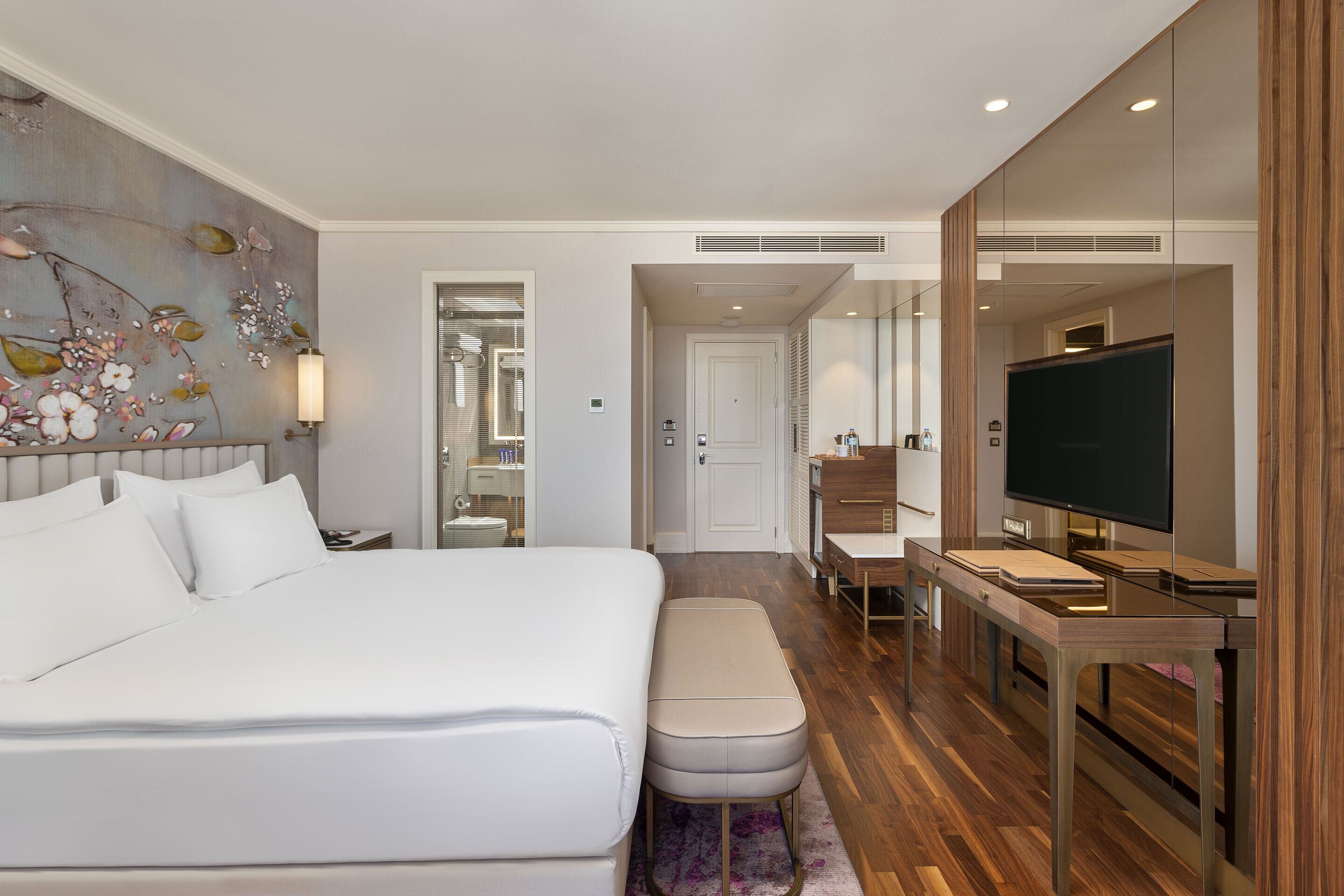 Quarto de hotel moderno com uma cama grande, uma escrivaninha, televisão e banheiro visível por uma porta aberta.