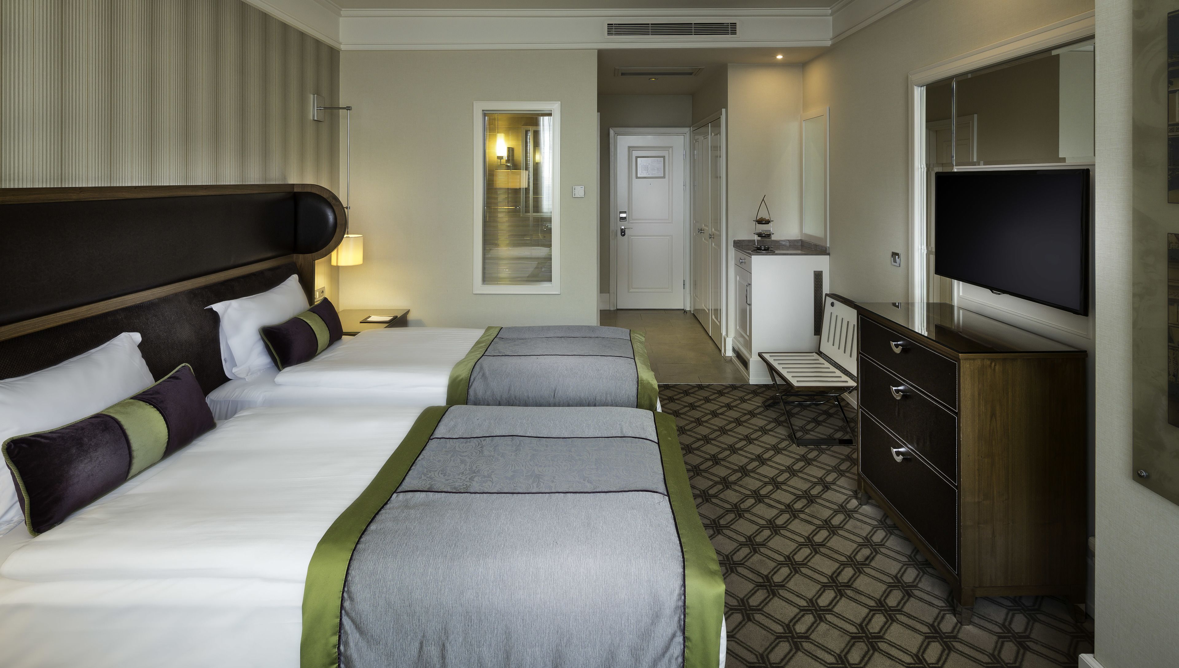 Habitación de hotel con dos camas, un televisor, un aparador y una puerta que da a un pasillo.