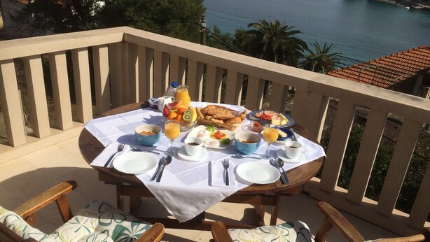 Ein Esstisch ist auf einem Balkon gedeckt, mit Blick auf eine Küstenlandschaft, das Meer und Boote.