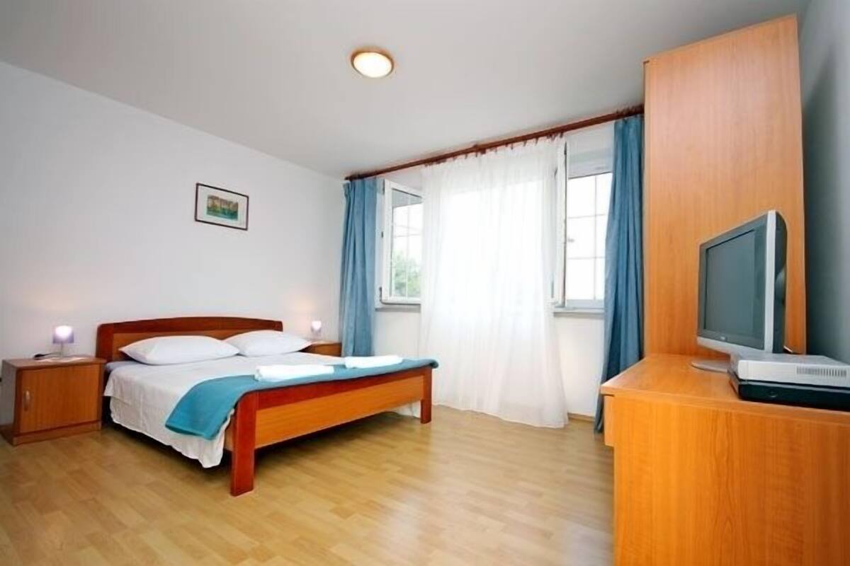 A bedroom with a wooden bed, a nightstand, a TV on a wooden cabinet, and a window with curtains.