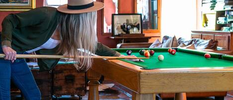A person playing pool in a room with a green pool table, a wooden cabinet, and a cowhide rug.