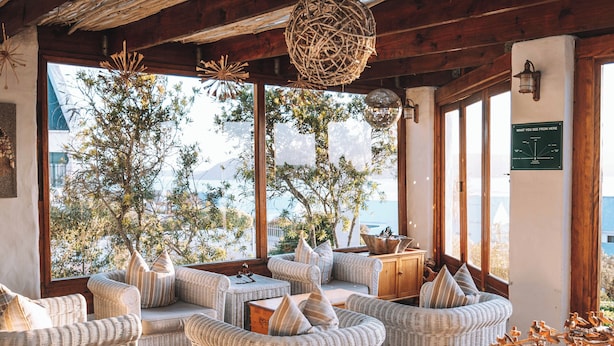 A cozy lounge area with wicker furniture, large windows offering a view of the outdoors, and a decorative hanging light fixture. - The Farmhouse Hotel (Langebaan)