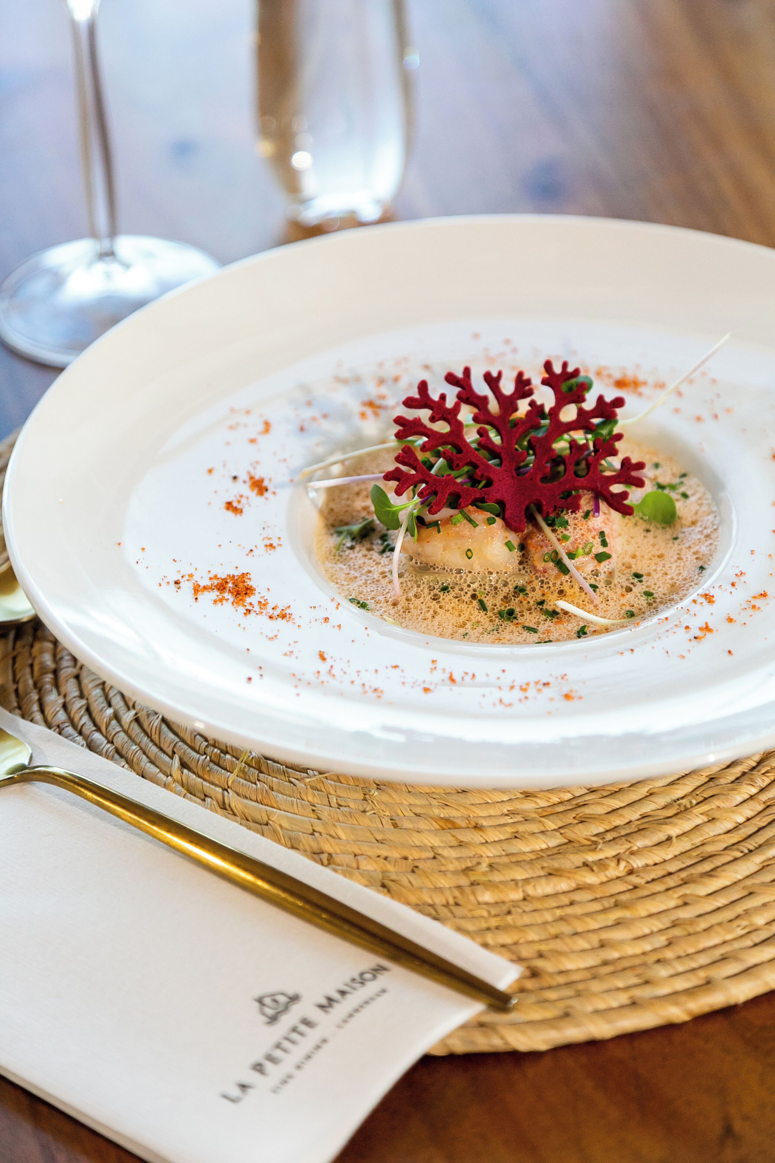 Un plato blanco con un plato de mariscos, adornado con coral rojo y hierbas verdes, sobre un camino de mesa tejido.