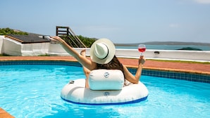 A person floating on an inflatable pool float in a clear blue pool with a view of the sea and a wooden deck in the background. - The Farmhouse Hotel (Langebaan)