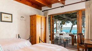 A room with two beds, wooden furniture, a view of the sea, and a balcony with a table and chairs. - The Farmhouse Hotel (Langebaan)