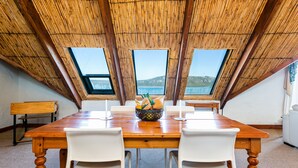 A dining area with a wooden table, white chairs, and a fruit bowl. There are three windows offering a view of the outdoors. - The Farmhouse Hotel (Langebaan)