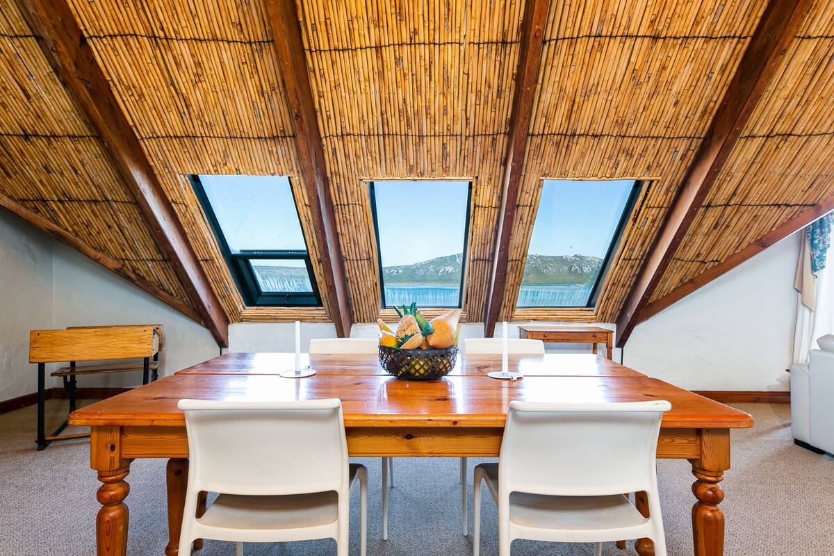A dining area with a wooden table, white chairs, and a fruit bowl. There are three windows offering a view of the outdoors.