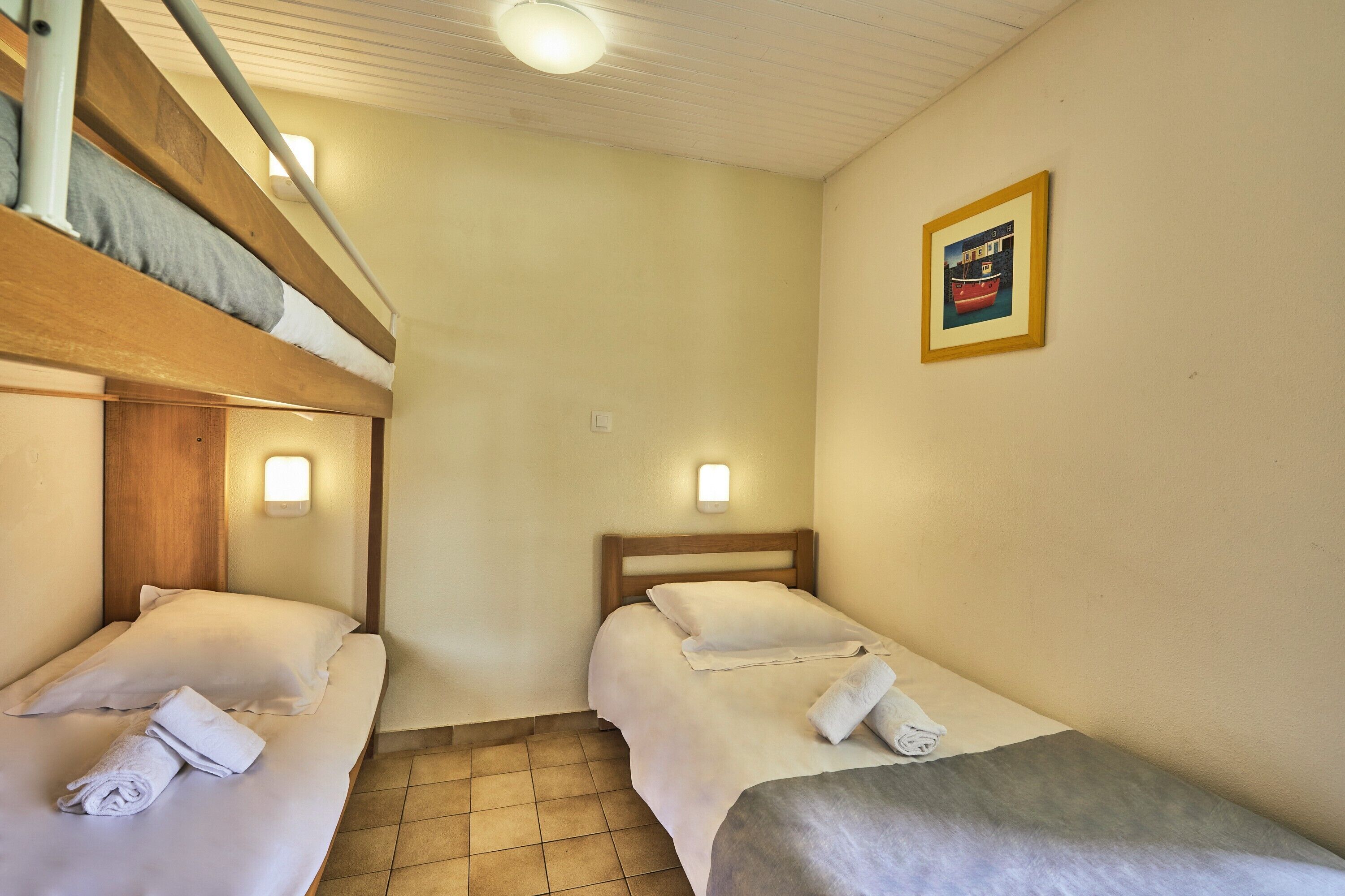Une chambre avec deux lits, chacun équipé d’un oreiller et d’une serviette enroulée. Un tableau encadré orne le mur, et un luminaire est fixé au plafond.