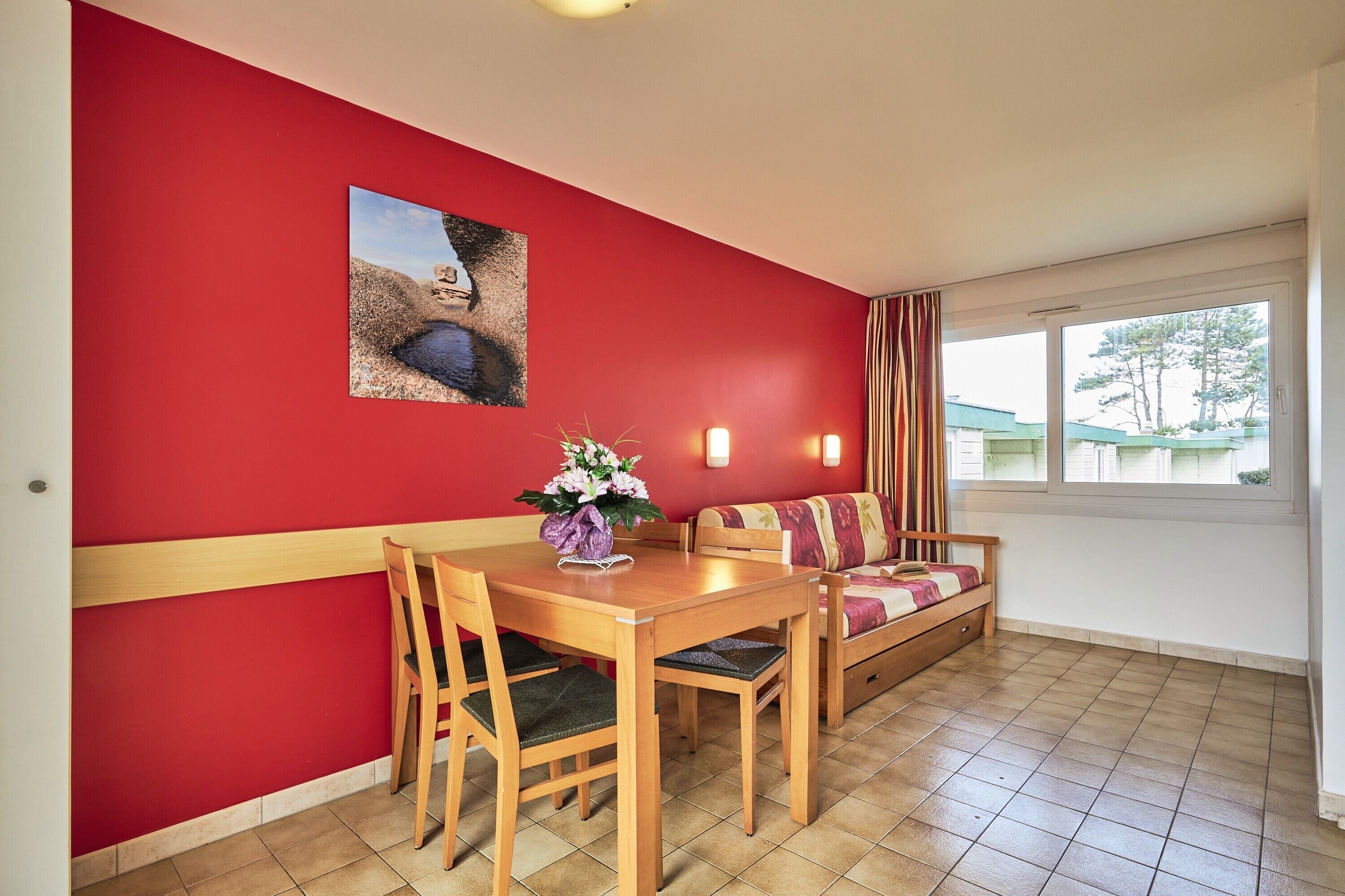 A dining area with a red wall, a wooden table, and chairs. There is a sofa with a cushion and a window with curtains.
