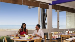 A dining area with a view of the beach, tables with chairs, and a buffet table with food and drinks.