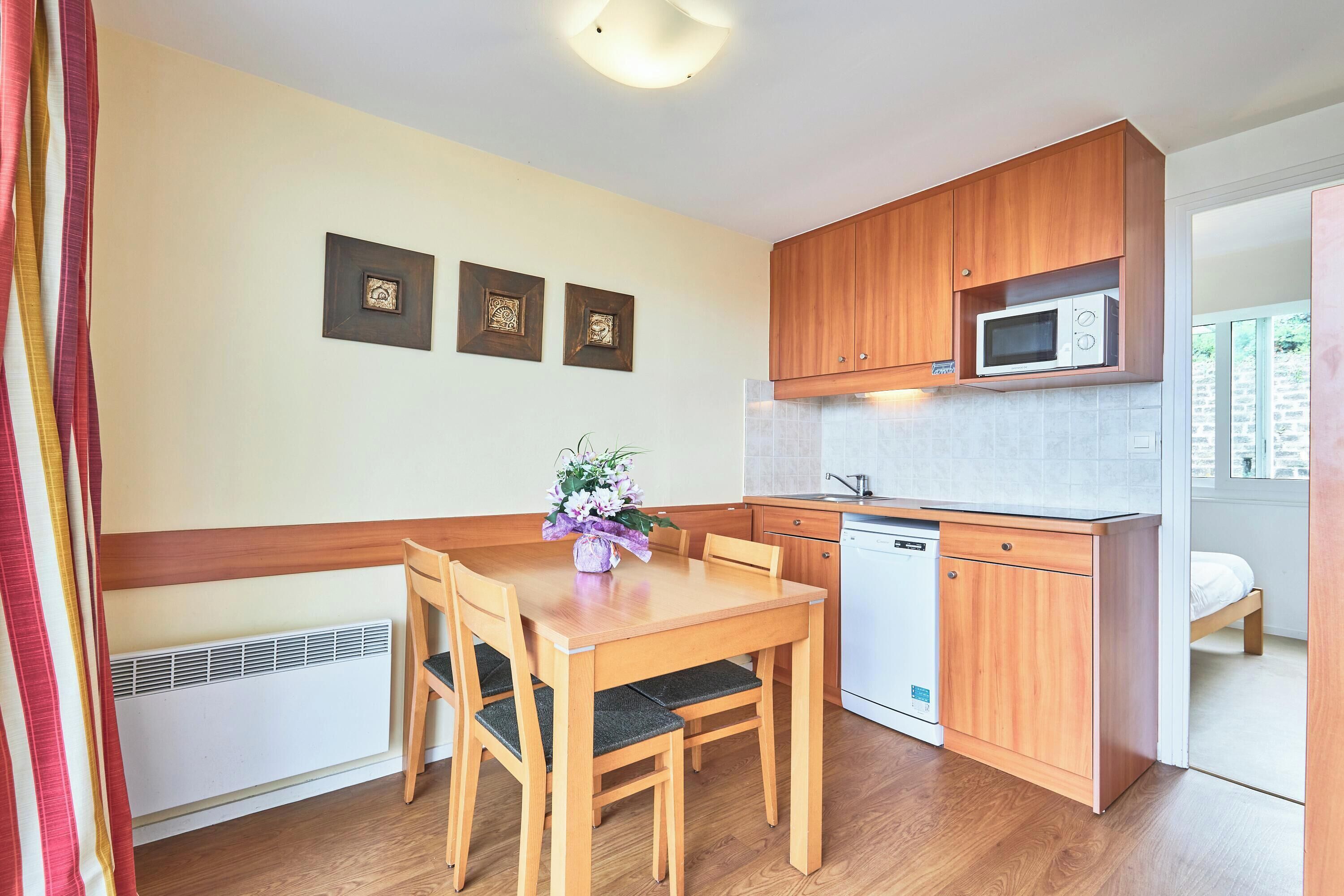 A compact kitchen with wooden cabinets, a microwave, a dishwasher, and a dining table with chairs. There are three framed pictures on the wall.