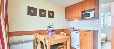 A compact kitchen with wooden cabinets, a microwave, a dishwasher, and a dining table with chairs. There are three framed pictures on the wall.