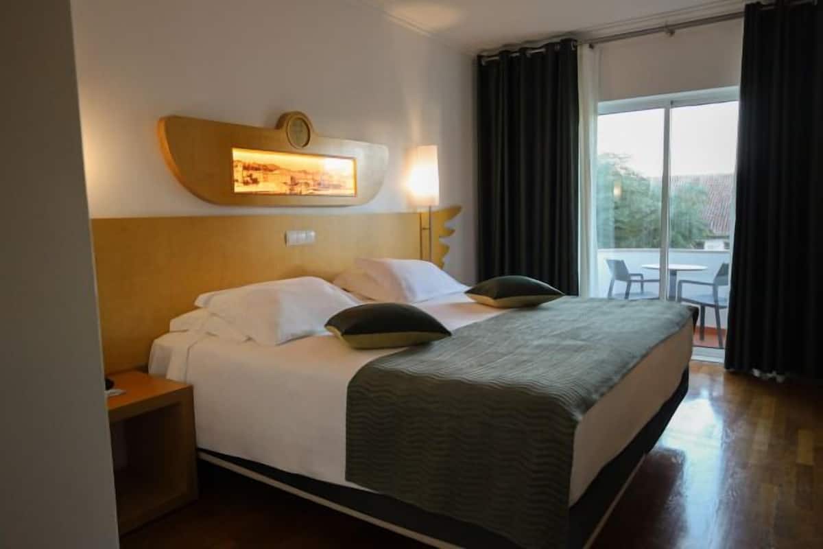 A hotel room with a bed, a nightstand, a wall-mounted lamp, and a view of the outdoors.