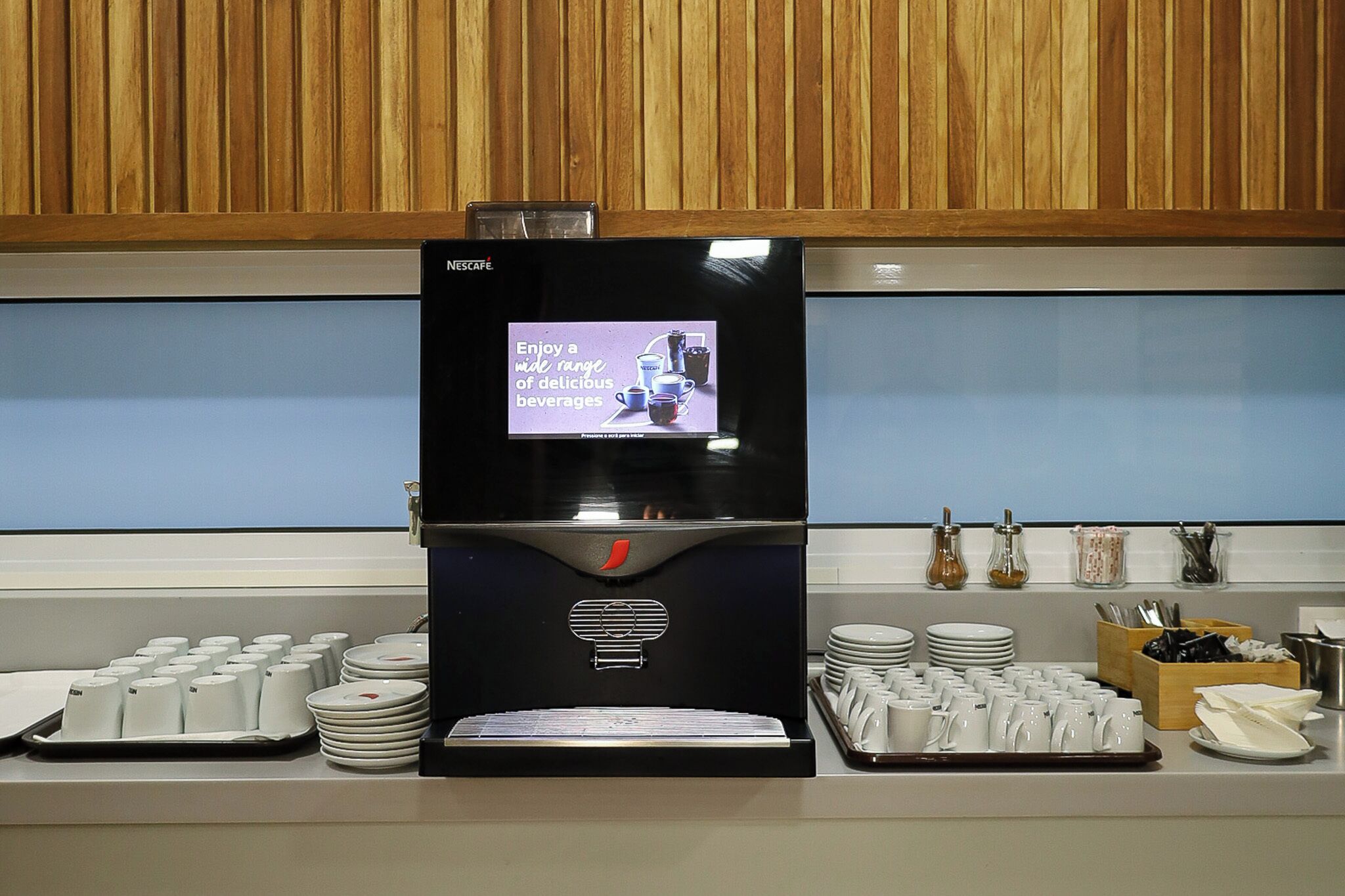 A Nescafe coffee machine with a digital display, surrounded by stacks of white plates and cups, and a tray of coffee mugs.