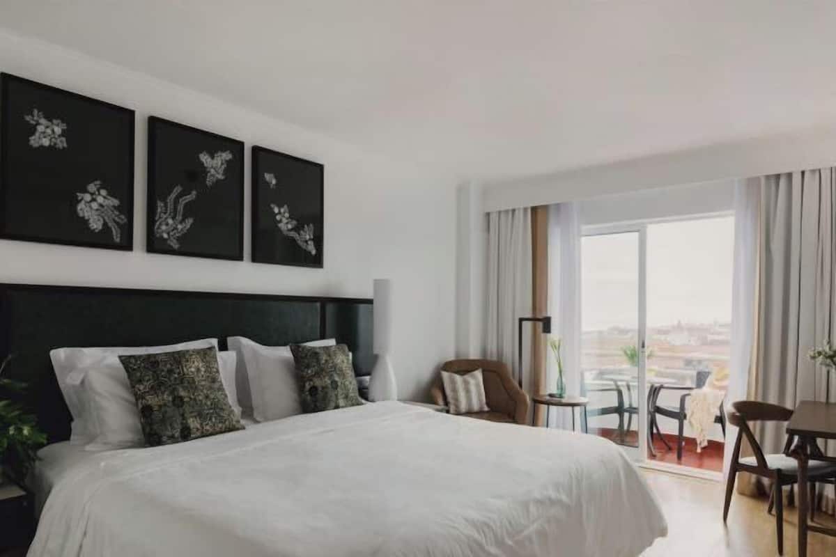 A hotel room with a large bed, a view of the city, and a balcony with a table and chairs.