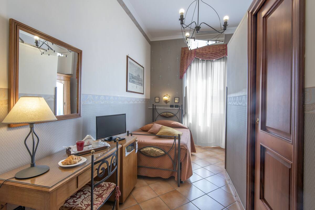 Una habitación de hotel con cama, escritorio con lámpara, televisor y ventana con cortinas.