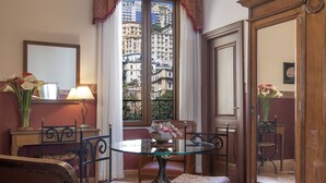 A room with a round glass table, two chairs, a bed, a desk with a lamp, and a window with a view of buildings. - Pinto Storey Hotel (Naples)