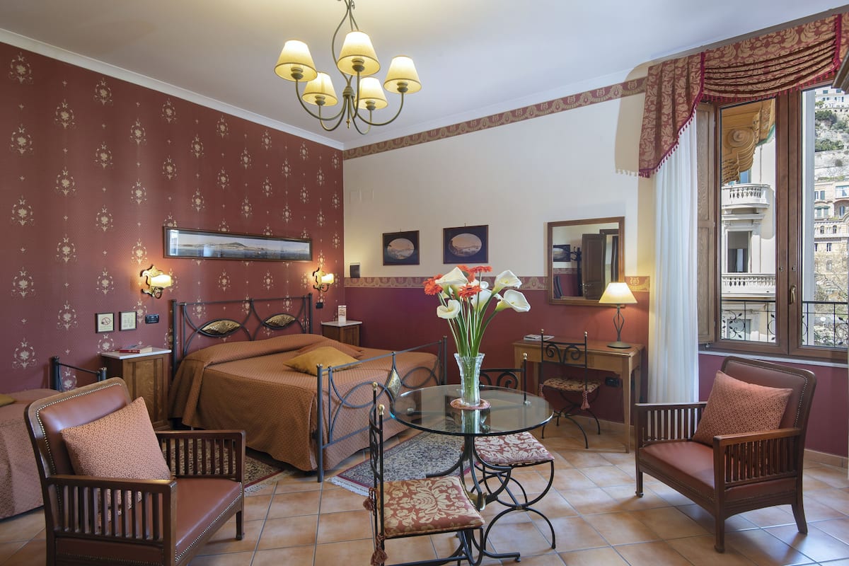 Habitación de hotel con cama, dos sillas, una mesa con un florero, un escritorio con una lámpara y una ventana con vistas a edificios.