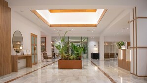 Un hall d’entrée spacieux avec un plafond haut, un grand bac à plantes au centre, et un comptoir de réception.