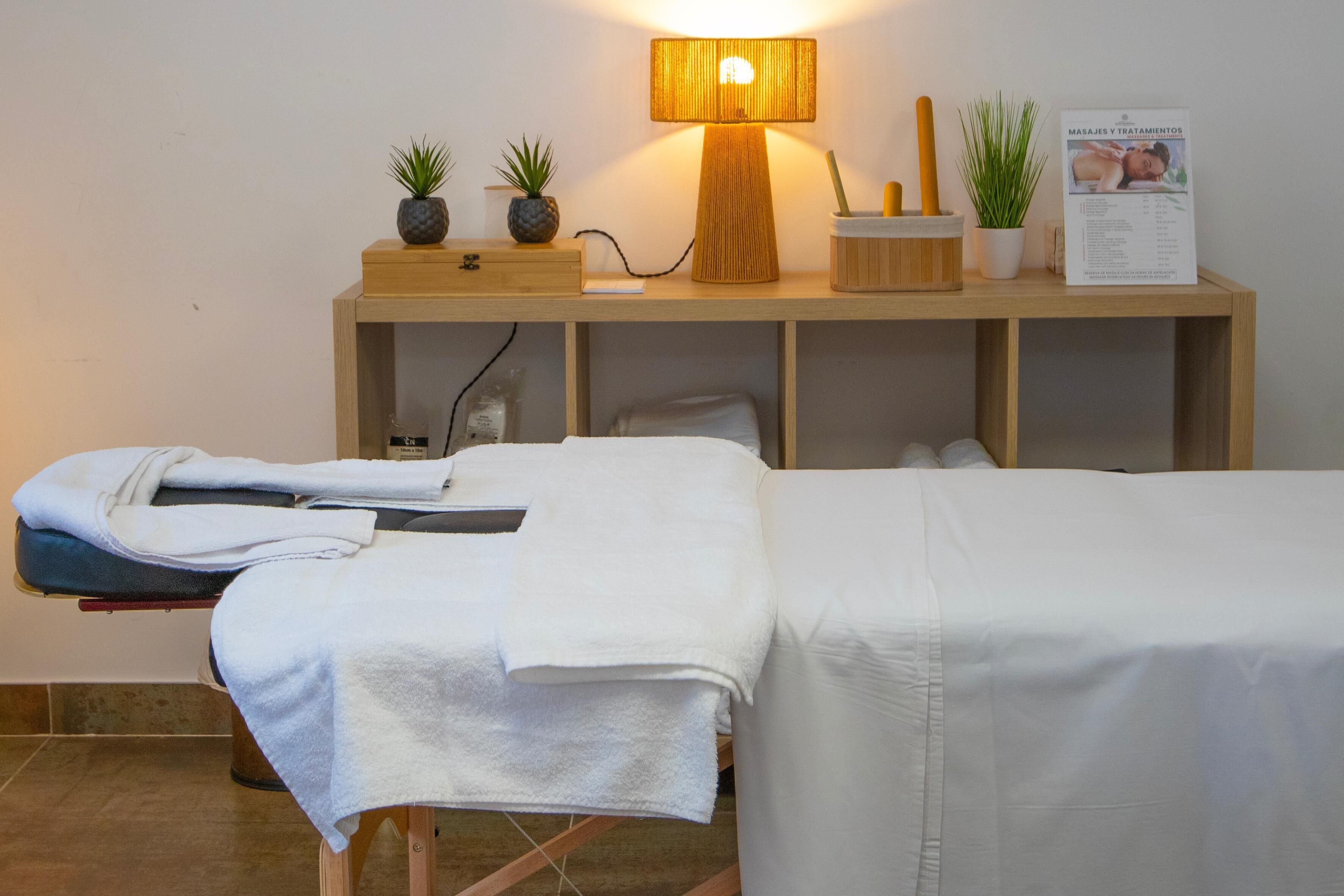 A massage therapy room with two massage tables, a wooden cabinet with plants, a lamp, and a poster on the wall.