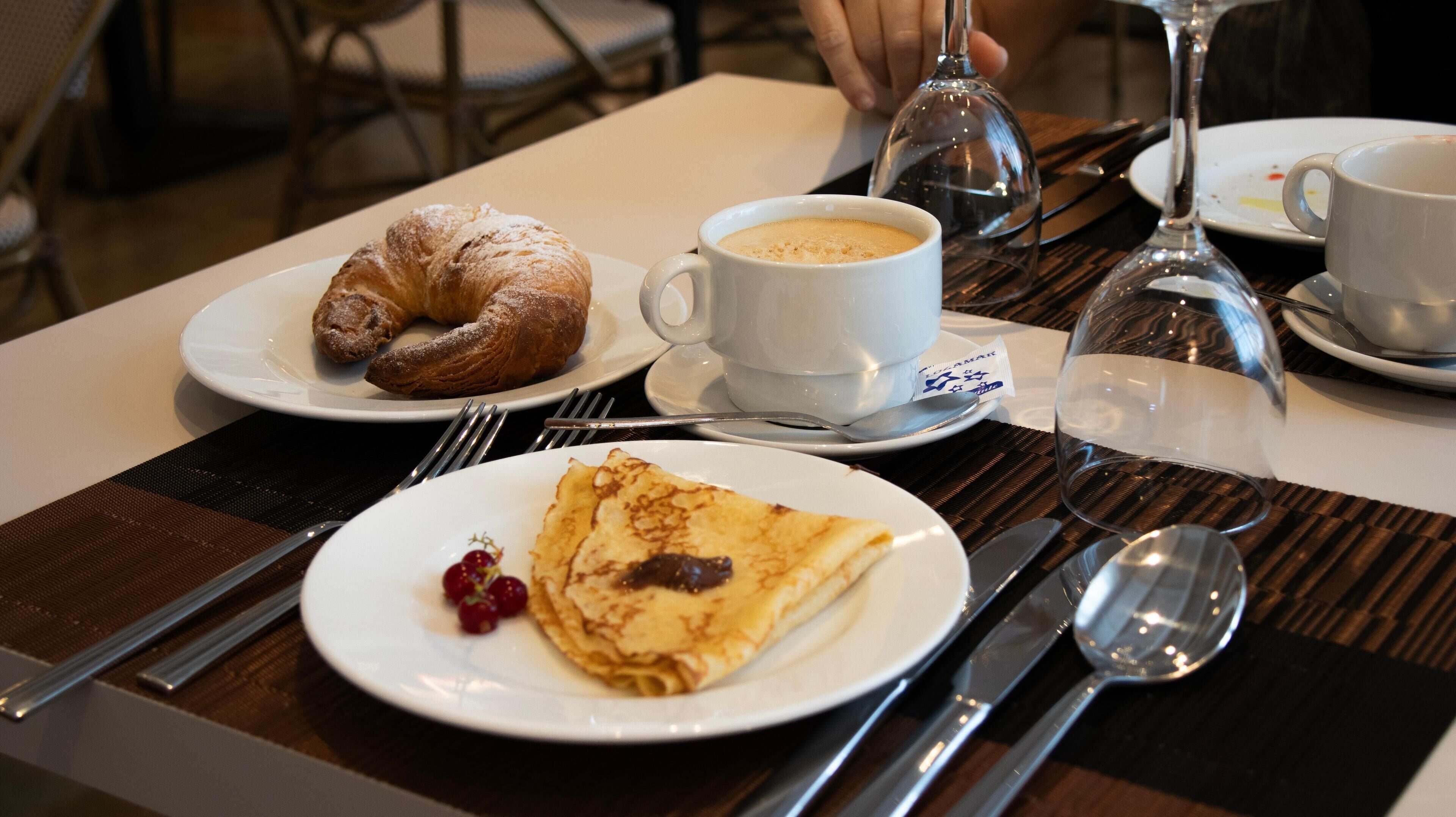 A dining table with plates of food, including a croissant, a cup of coffee, and a crepe with berries.