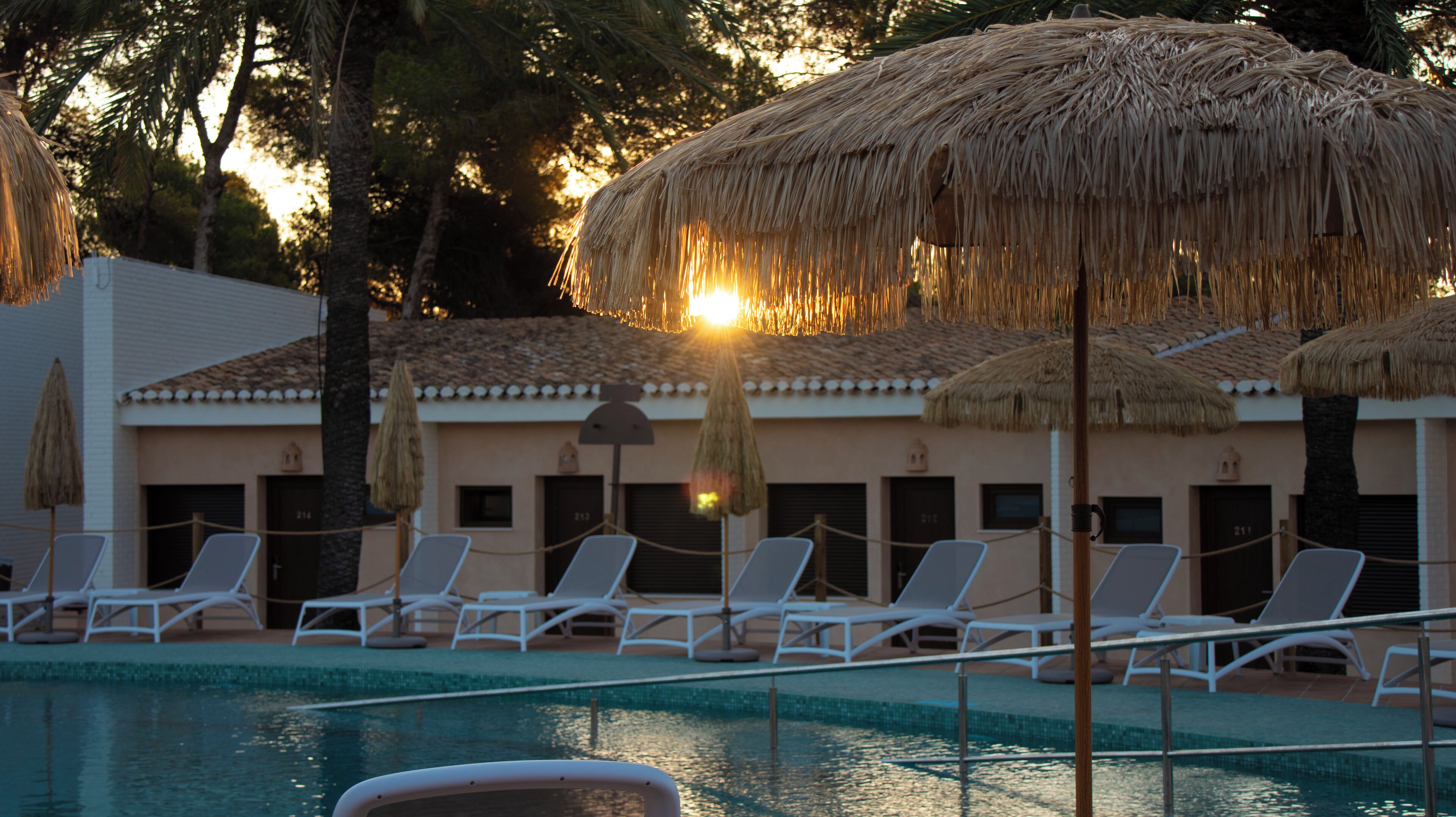 A poolside area with lounge chairs and thatched umbrellas, surrounded by a building with a tiled roof.