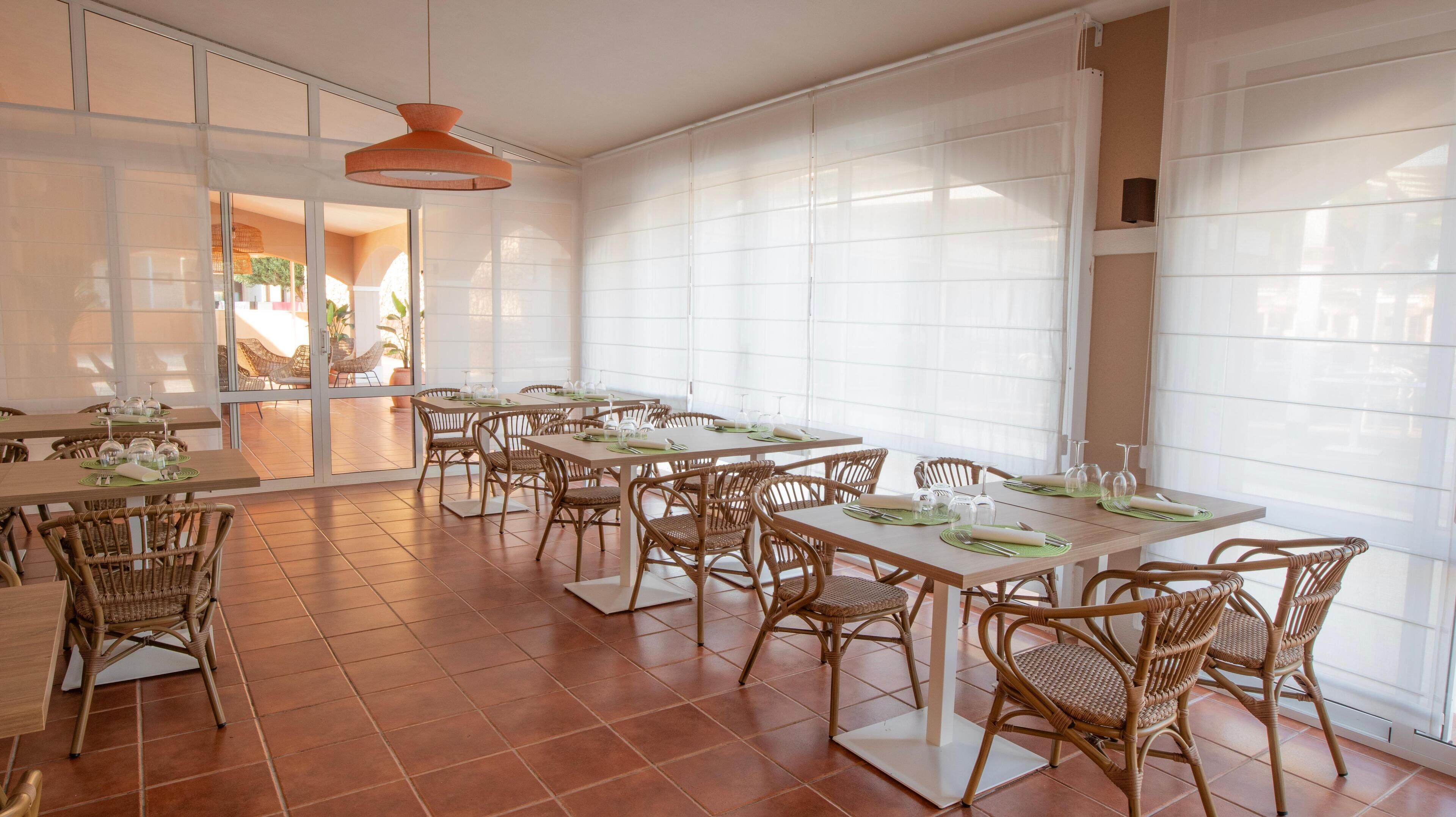 A dining area with multiple tables and wicker chairs, large windows, and a hanging light fixture.