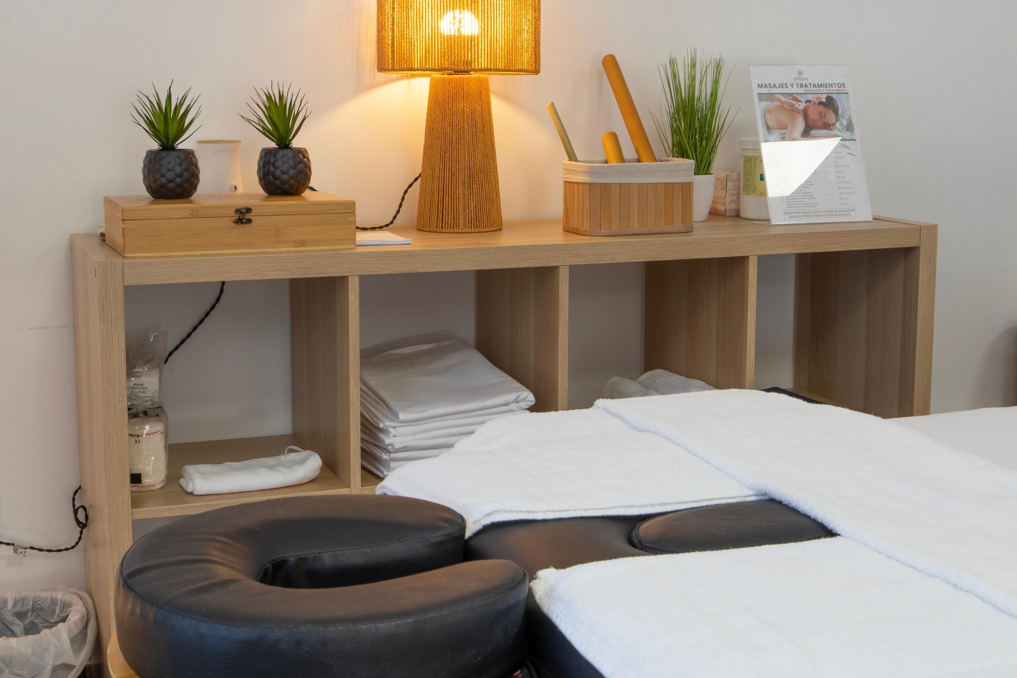 A neatly arranged room with a bed, a massage chair, a small table with plants, a lamp, and a sign about massages and treatments.