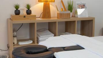 A neatly arranged room with a bed, a massage chair, a small table with plants, a lamp, and a sign about massages and treatments.