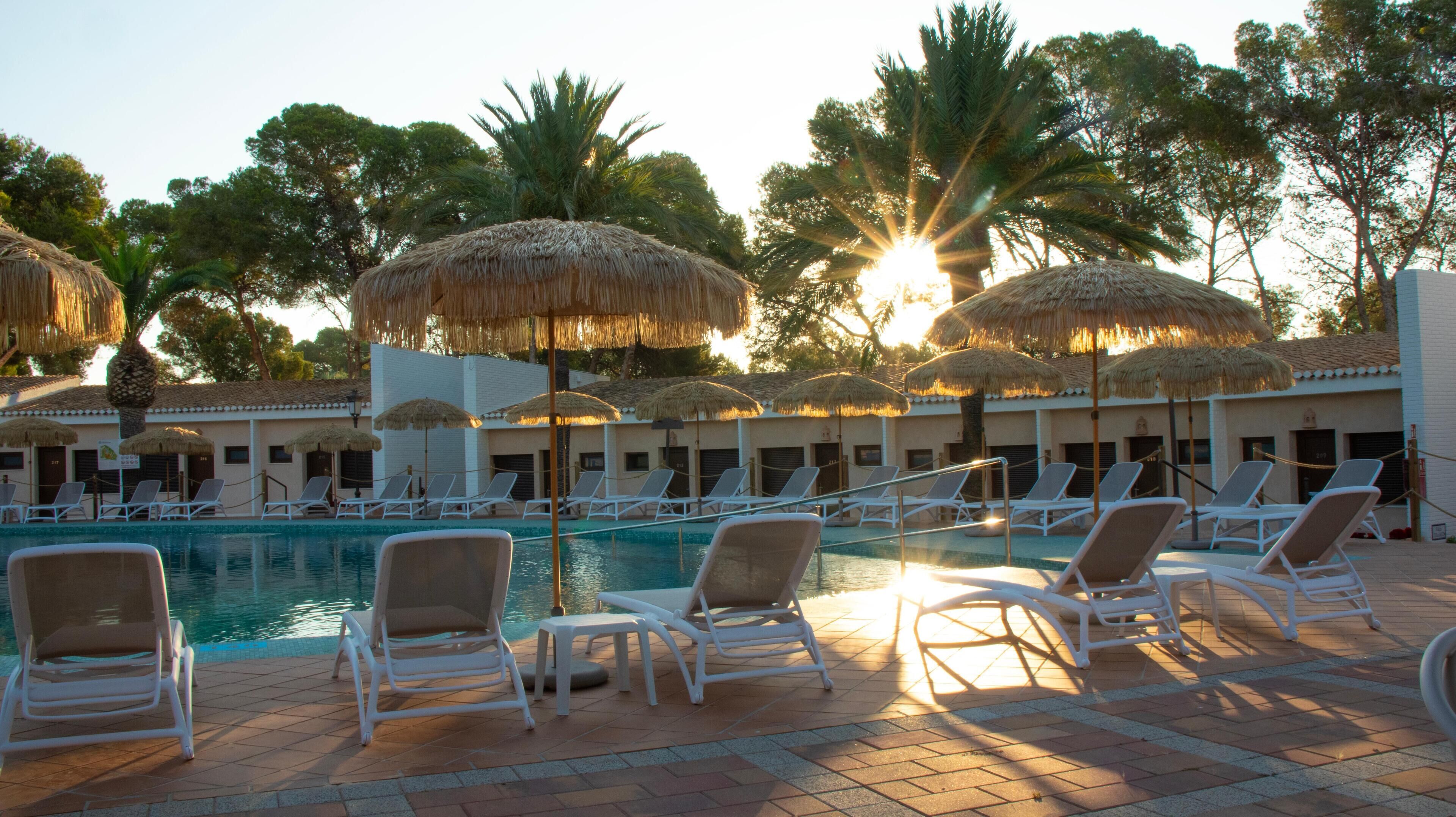 A poolside area with lounge chairs and thatched umbrellas, surrounded by palm trees and a building with a tiled roof.
