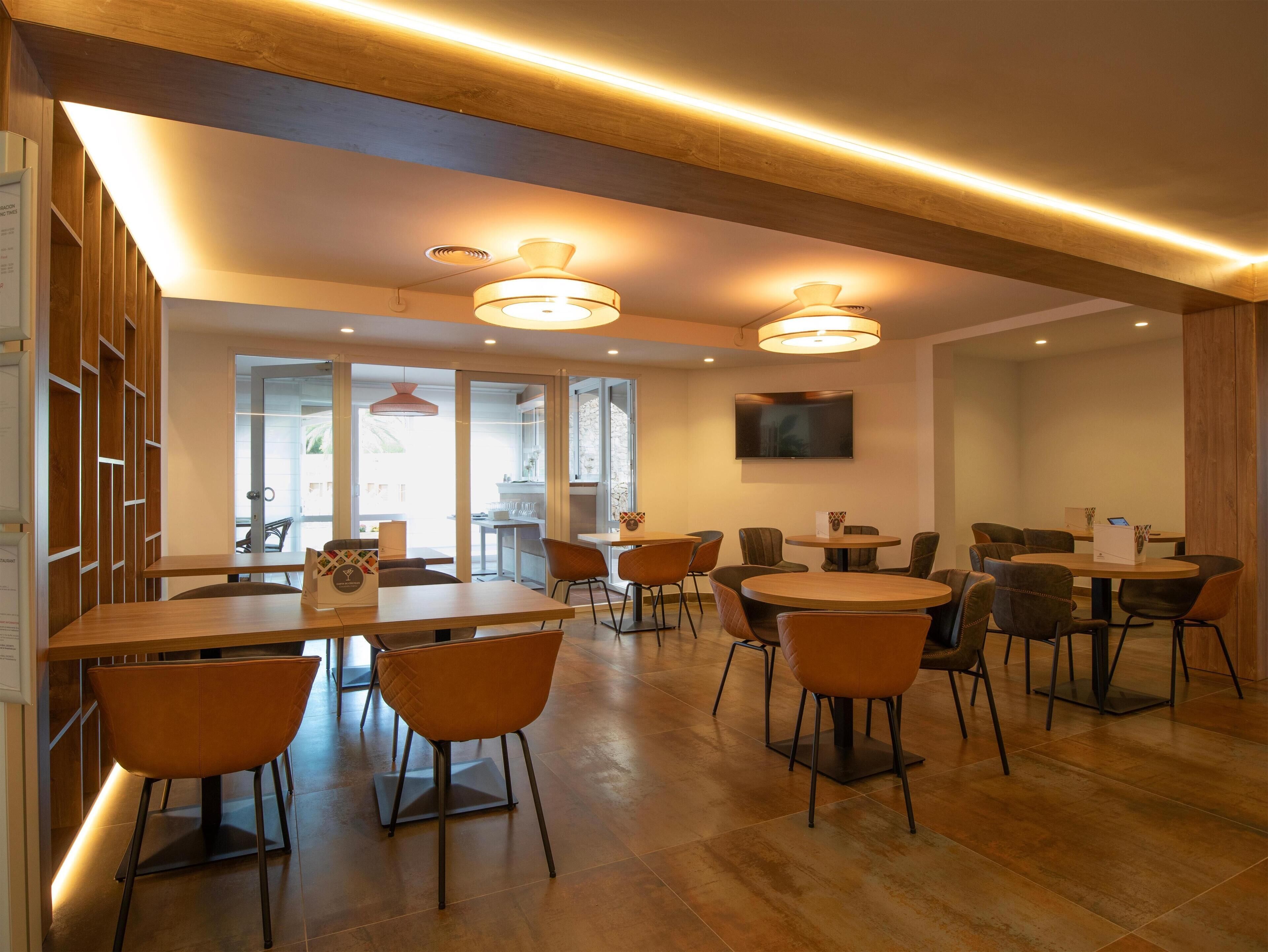 A modern dining area with wooden tables and chairs, recessed ceiling lighting, and a flat-screen TV on the wall.