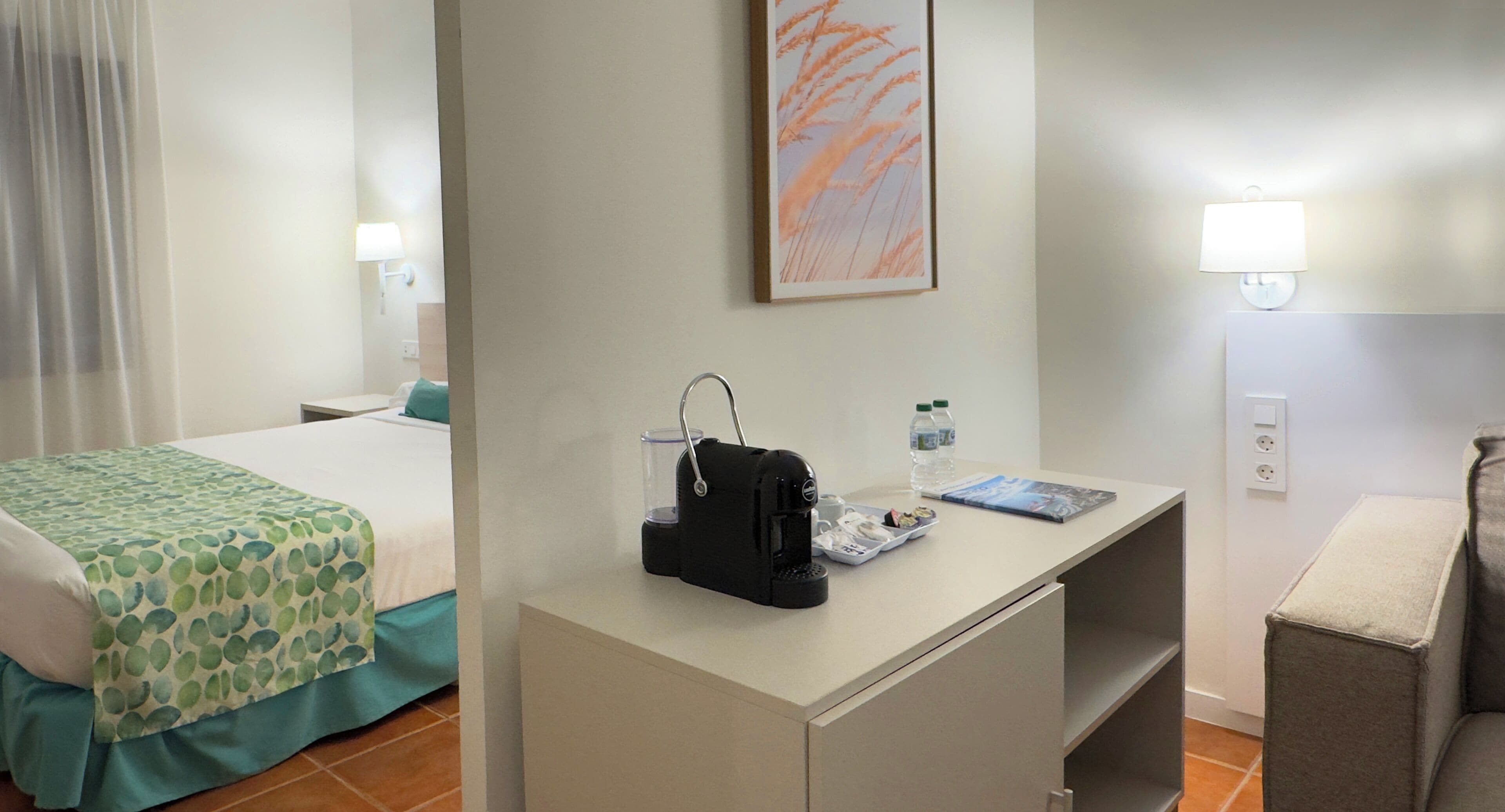 A hotel room with a bed, a desk with a lamp, a picture on the wall, and a chair.