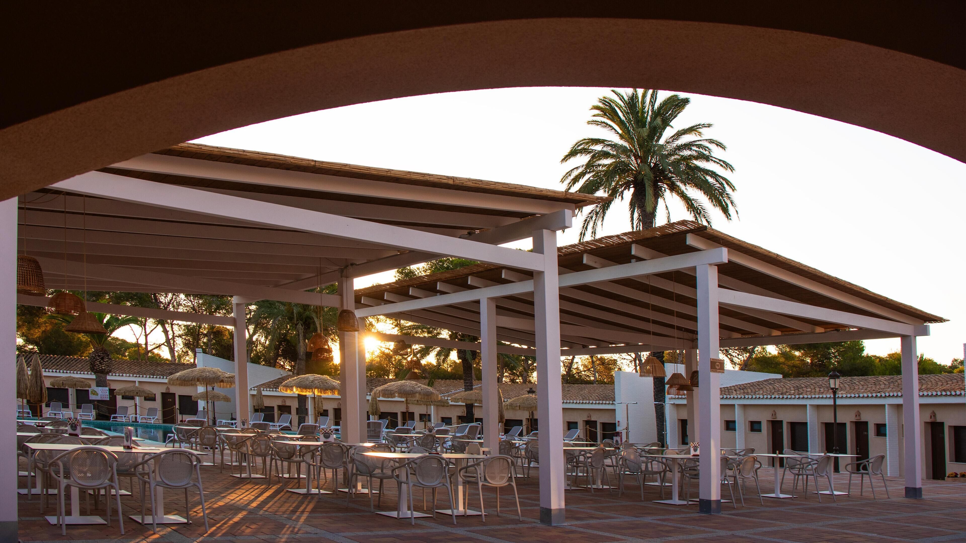 A covered outdoor seating area with tables and chairs, a pool, and palm trees.