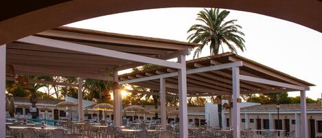 A covered outdoor seating area with tables and chairs, a pool, and palm trees.