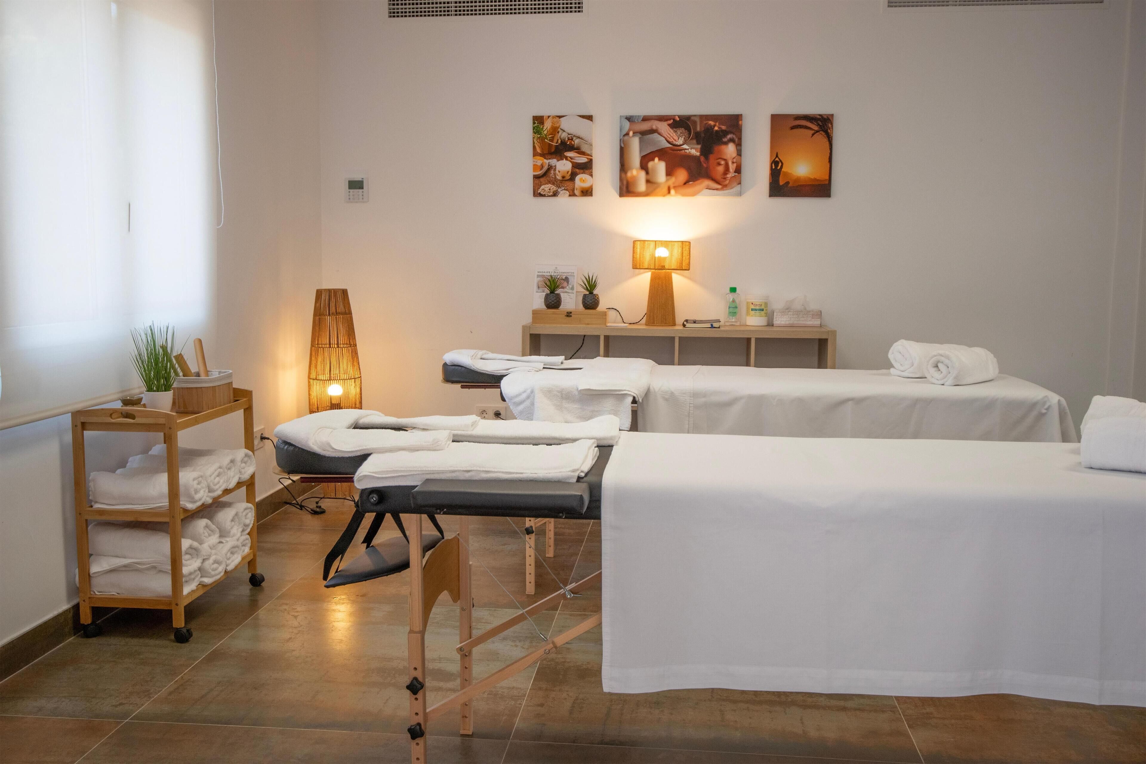 Un cuarto de spa con dos camillas, una mesa de masaje, un estante con toallas, una lámpara y decoración en la pared.