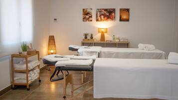 Un cuarto de spa con dos camillas, una mesa de masaje, un estante con toallas, una lámpara y decoración en la pared.