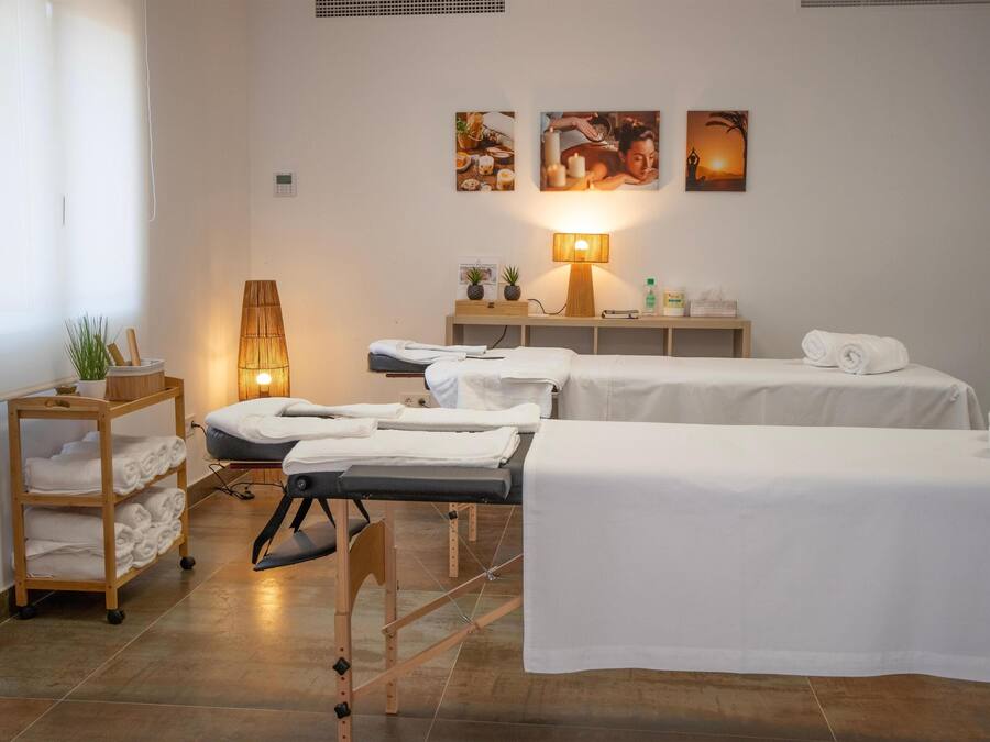 Une salle de spa équipée de deux tables de soins, d’une table de massage, d’une étagère avec des serviettes, d’une lampe et d’œuvres d’art murales.