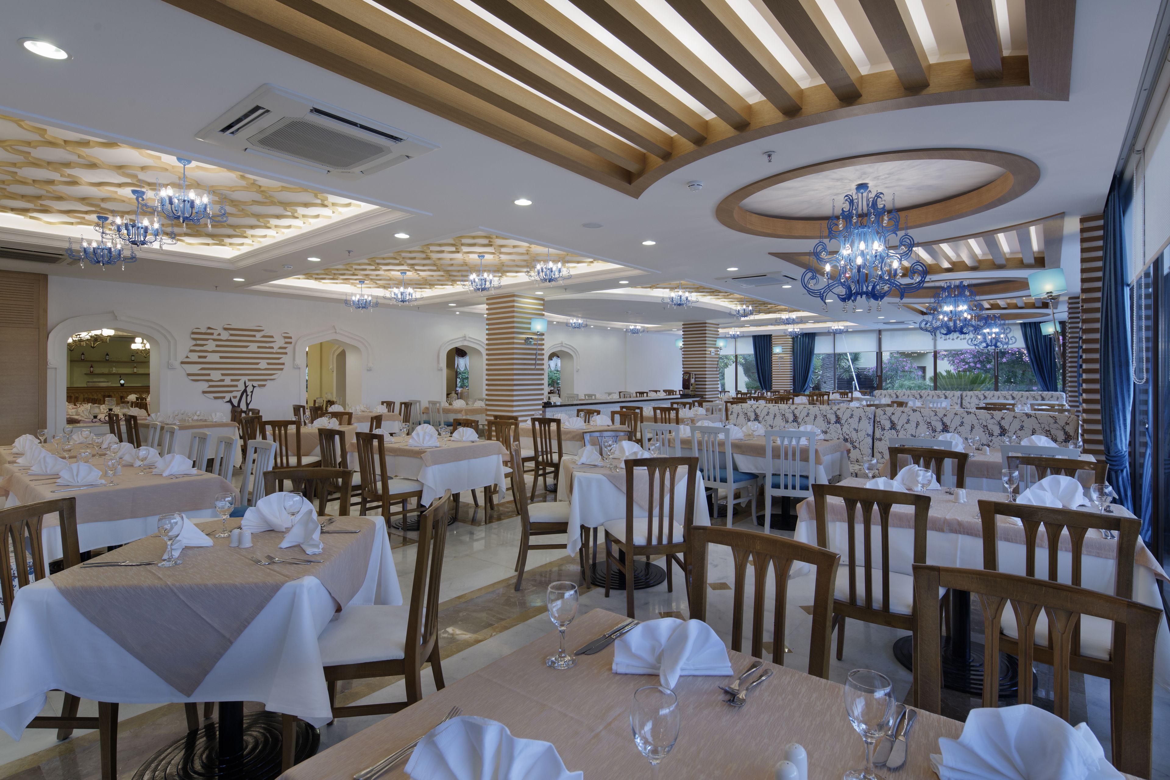A spacious dining area with multiple tables set for dining, elegant chandeliers, and a modern ceiling design.
