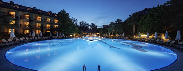 A large, circular pool with lounge chairs and umbrellas around it, surrounded by multi-story buildings with balconies and outdoor lighting.