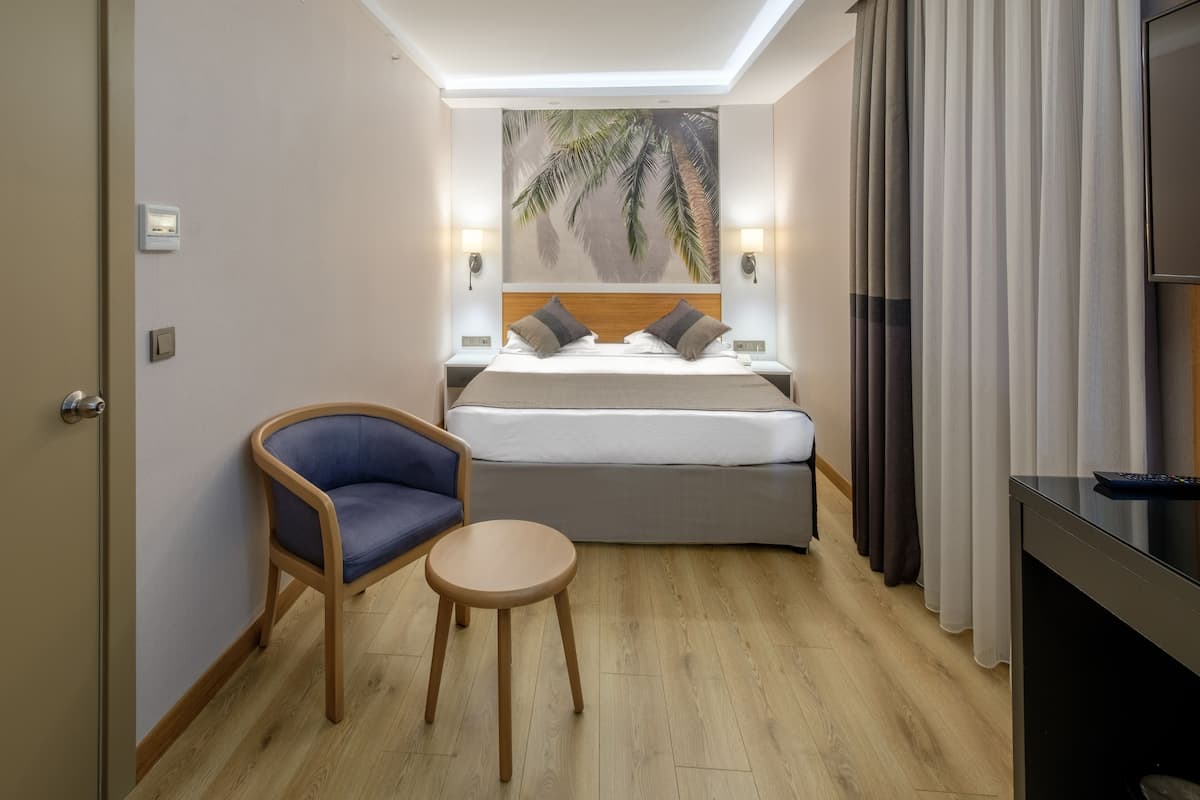 Ein Hotelzimmer mit Bett, Sessel, kleinem Tisch, Fernseher und einem Palmenbild an der Wand.