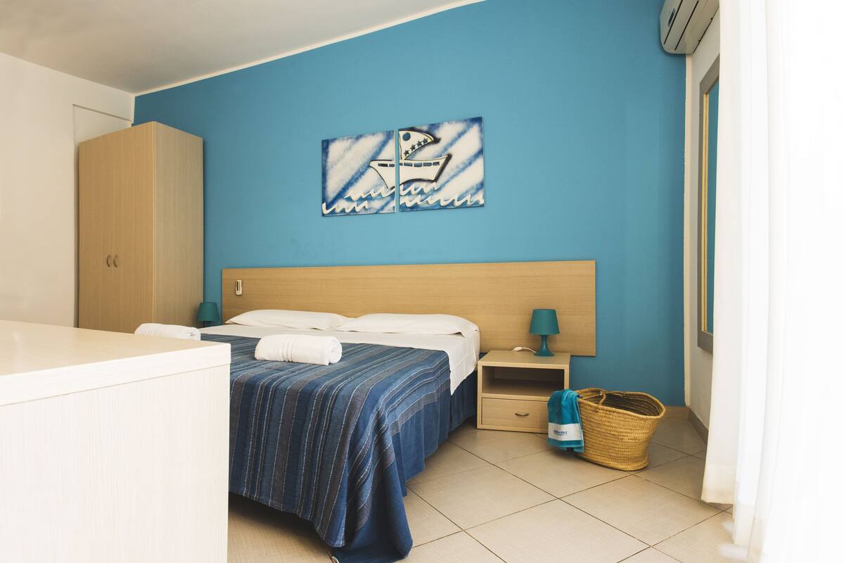 Une chambre d’hôtel comprenant un lit, une table de chevet, une armoire en bois, une œuvre d’art murale et un panier en osier.