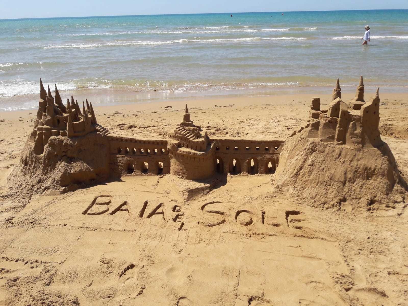 Um castelo de areia com a inscrição "BAJA SOLLE" numa praia, com o oceano ao fundo.