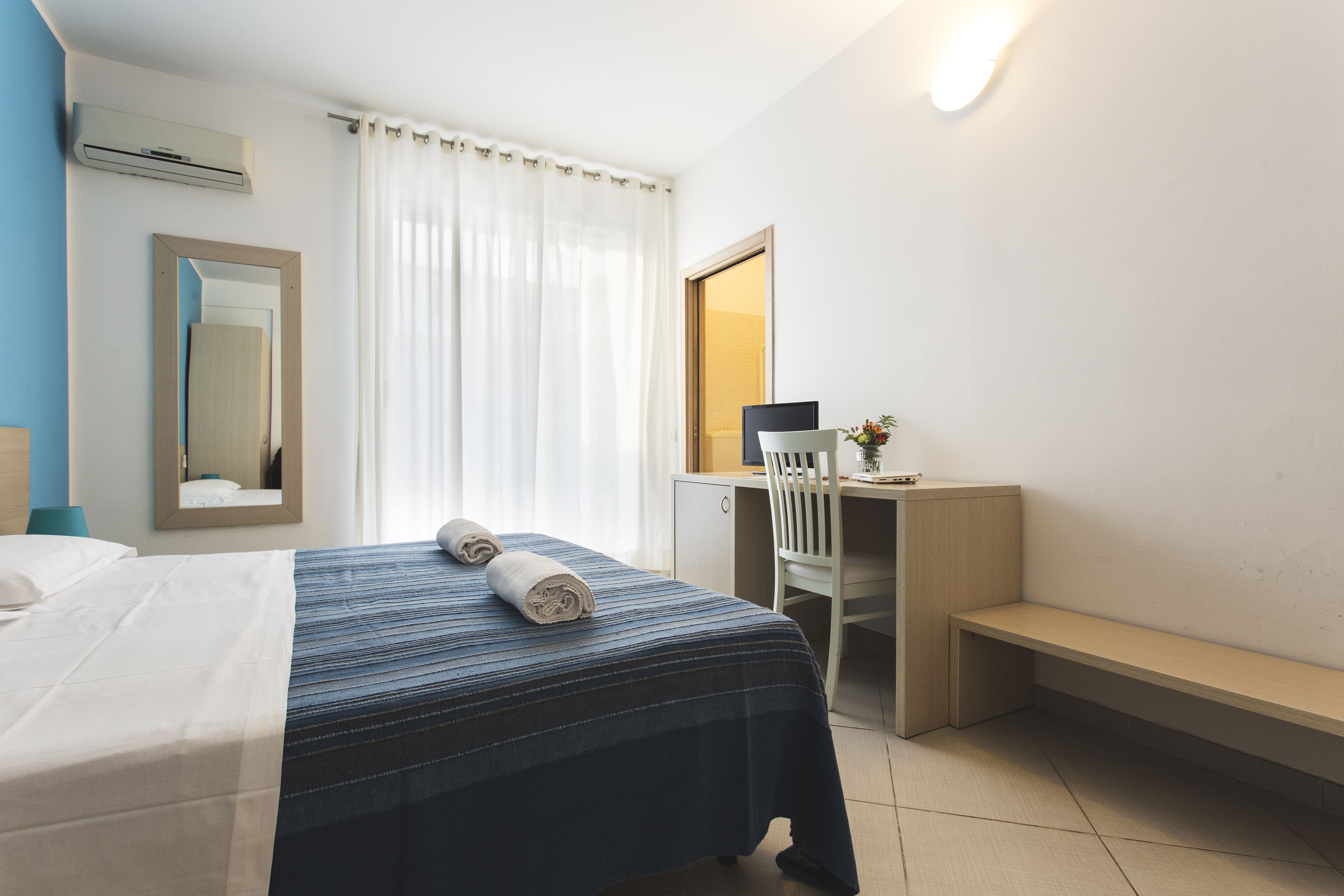 Quarto de hotel com cama, escrivaninha, cadeira, banco e espelho.